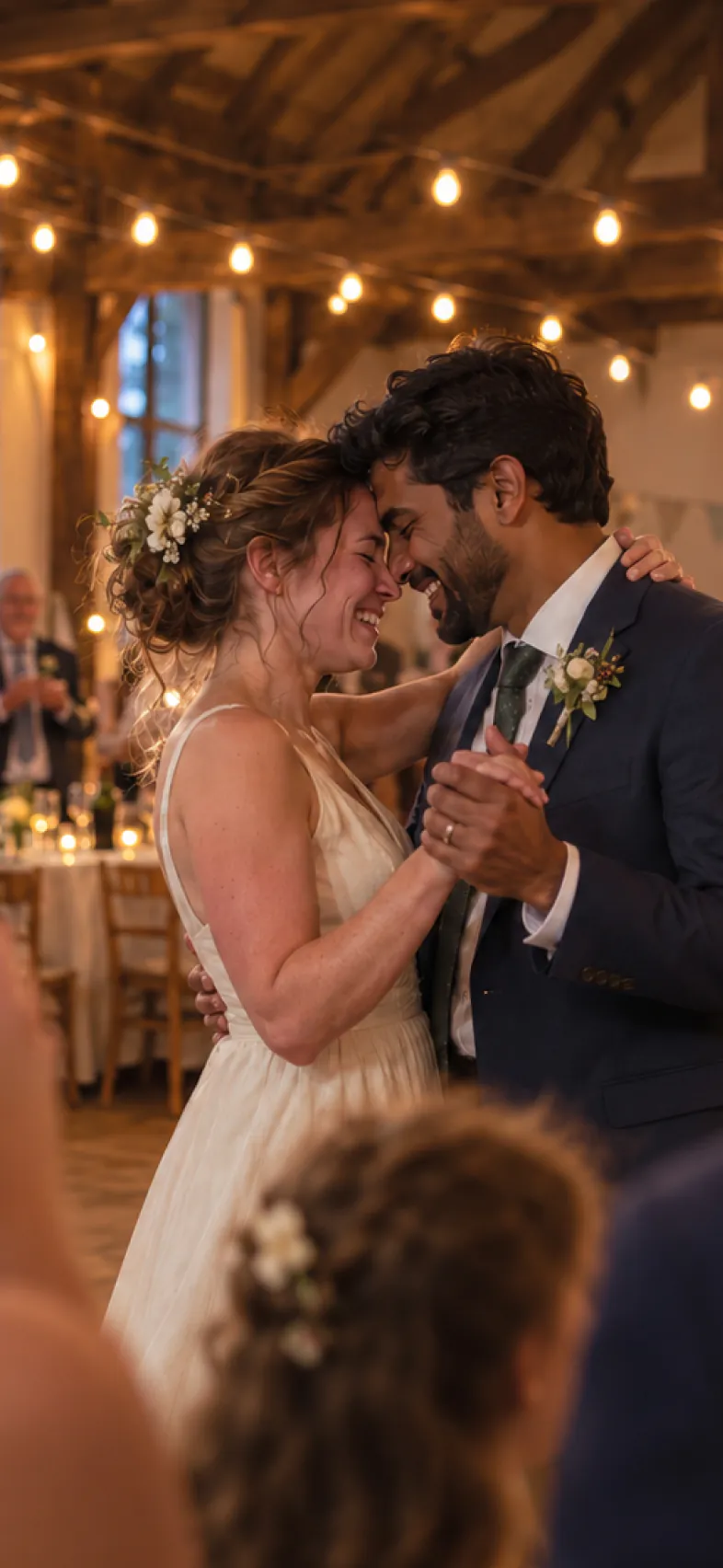 A bride and groom dancing cheek to cheek at their wedding in a barn hung with string lights